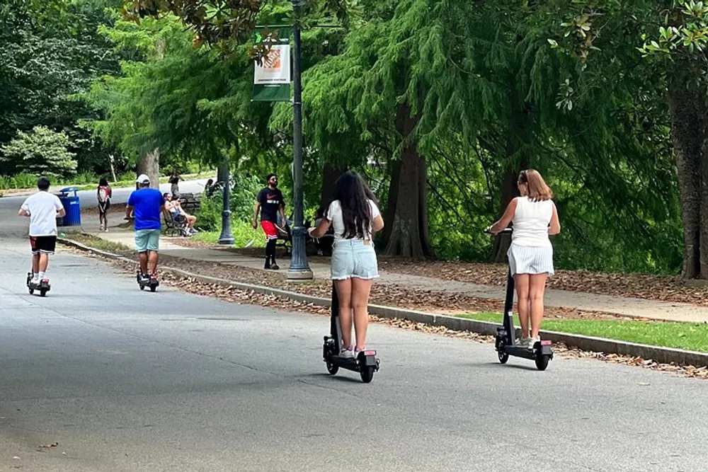 Two individuals are riding electric scooters along a park path with other park-goers visible in the background