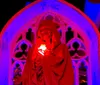 A person draped in a red robe stands before a gothic arch illuminated in red light cradling a glowing white object to their chest