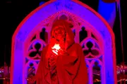 A person draped in a red robe stands before a gothic arch illuminated in red light, cradling a glowing white object to their chest.