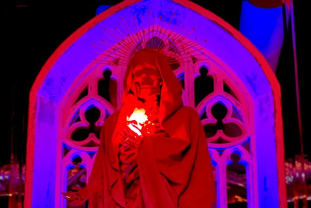 A person draped in a red robe stands before a gothic arch illuminated in red light cradling a glowing white object to their chest