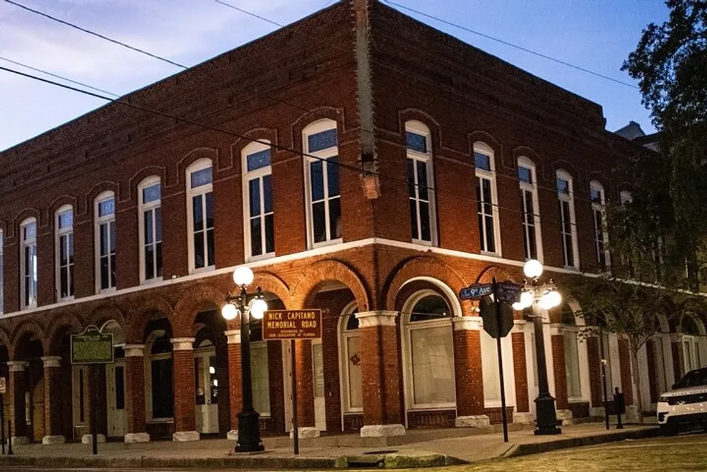 The image shows a two-story brick corner building with arched windows and a sign for Vice Capitano Memorial Road taken at dusk with street lighting illuminating the facade and a single vehicle parked on the side