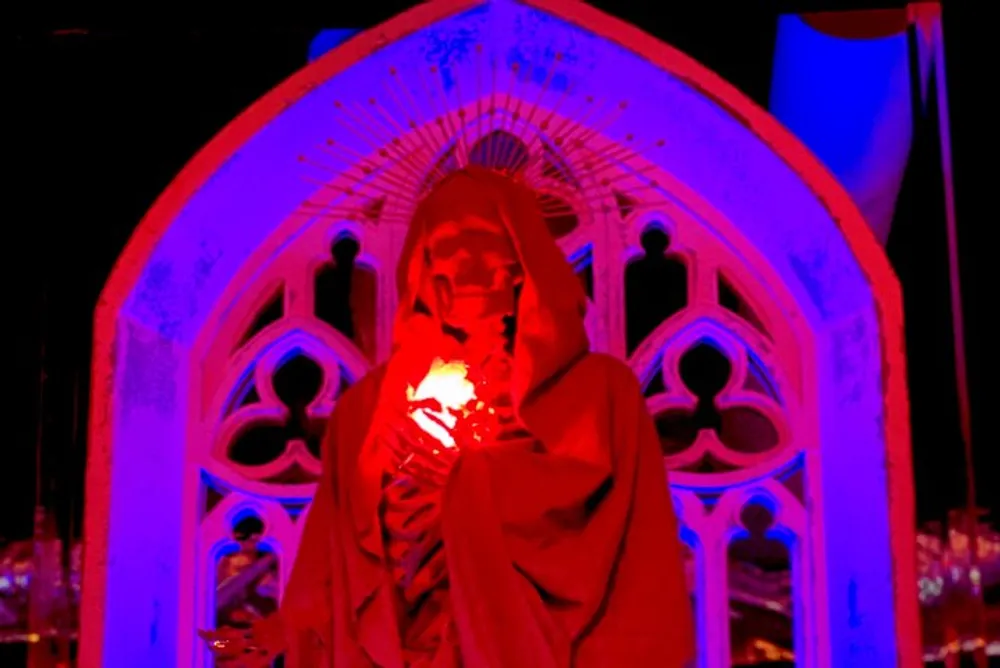 A person draped in red fabric stands before a gothic archway illuminated in eerie red light and holding a glowing object to their face