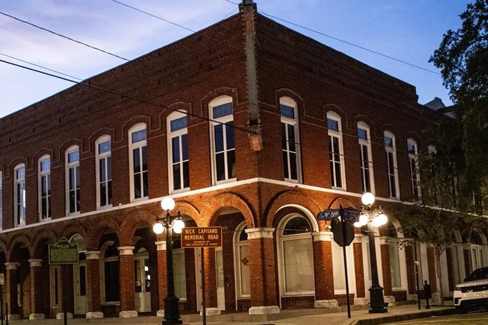 The image captures a two-story brick corner building with arched openings on the ground floor illuminated by streetlights at dusk or dawn