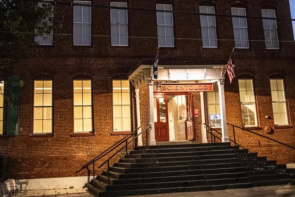 The image shows the exterior of a brick building at dusk with a lit-up entrance sign that reads Scientology an American flag and steps leading up to the door
