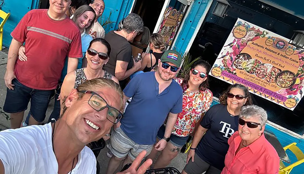 A group of people are posing for a selfie in front of a food truck serving Pakistani and Indian cuisine