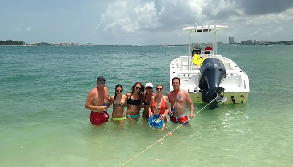 A group of people is enjoying a sunny day in shallow waters beside a boat with some holding beverages and the coastline visible in the background