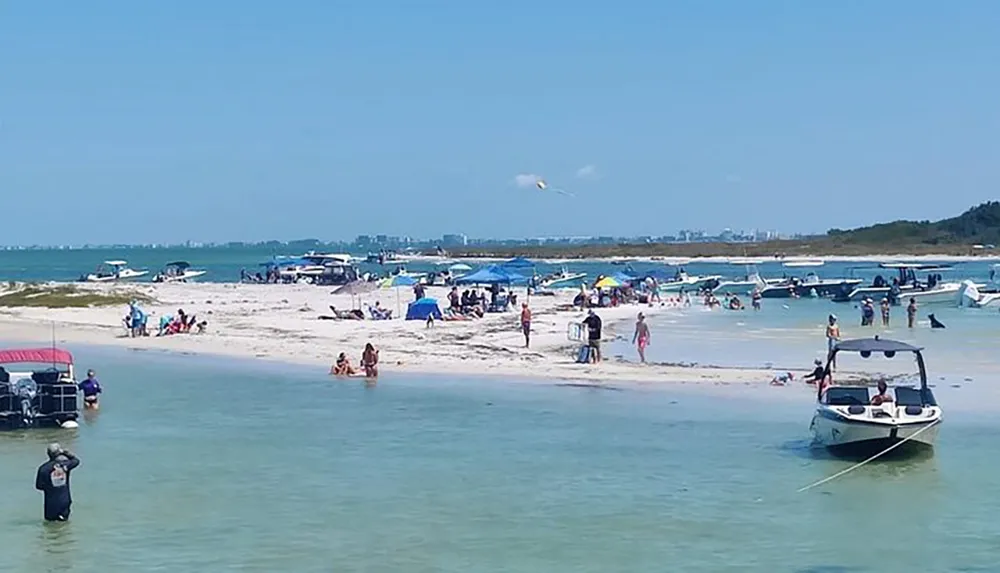 The image shows a bustling beach scene with people enjoying the sun sand and water alongside moored boats and beach umbrellas