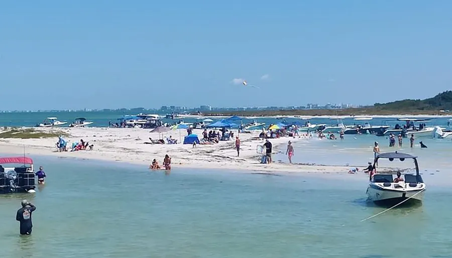The image shows a bustling beach scene with people enjoying the sun, sand, and water, alongside moored boats and beach umbrellas.