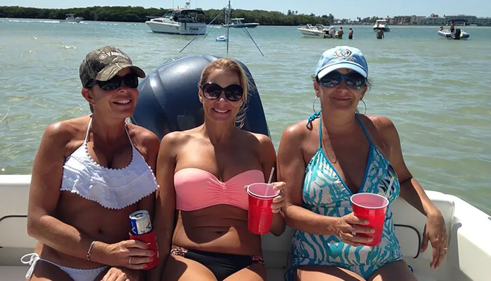 Three people are smiling on a boat with drinks in their hands on a sunny day with other boats and water in the background