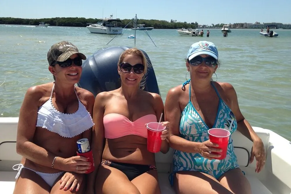 Three individuals are seated smiling on a boat holding beverages with the backdrop of a sunny day and other boats on the water