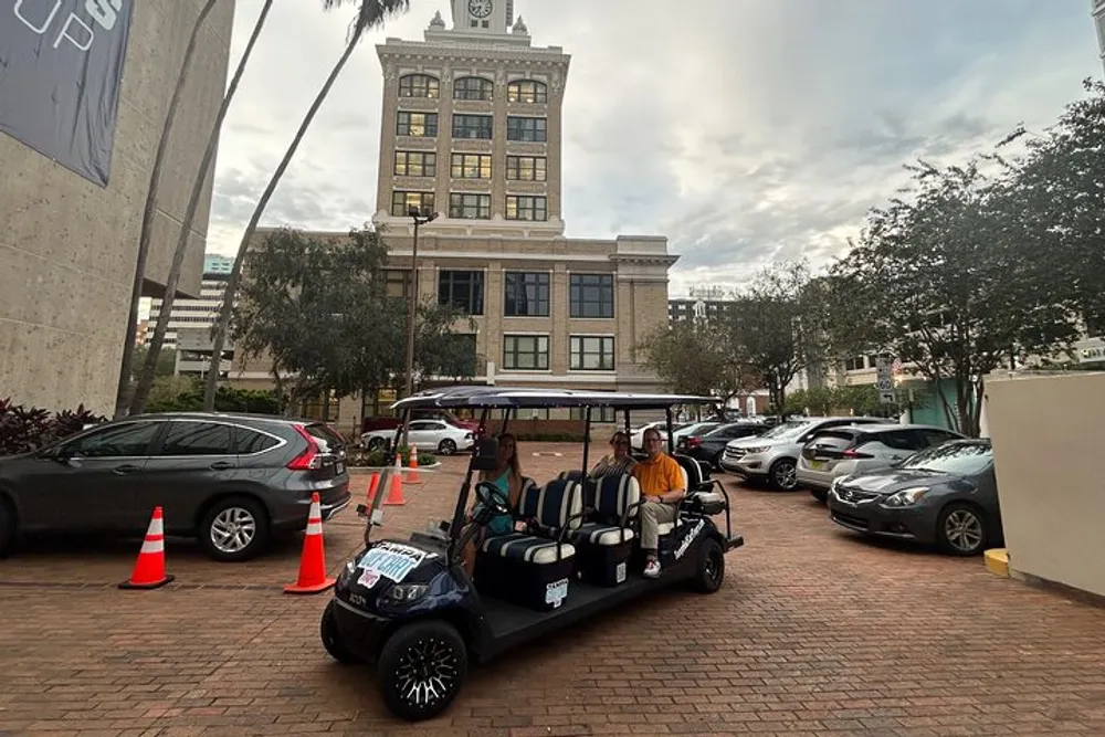 A golf cart with passengers is parked on a brick-paved area near a valet stand in front of a historic building with a clock tower