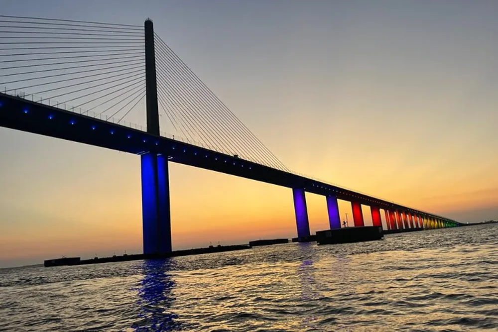 A cable-stayed bridge illuminated with blue lights spans over water against a vibrant sunset sky gradating from yellow to blue