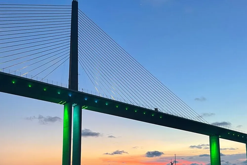 A cable-stayed bridge illuminated with green lights stands against a vibrant evening sky