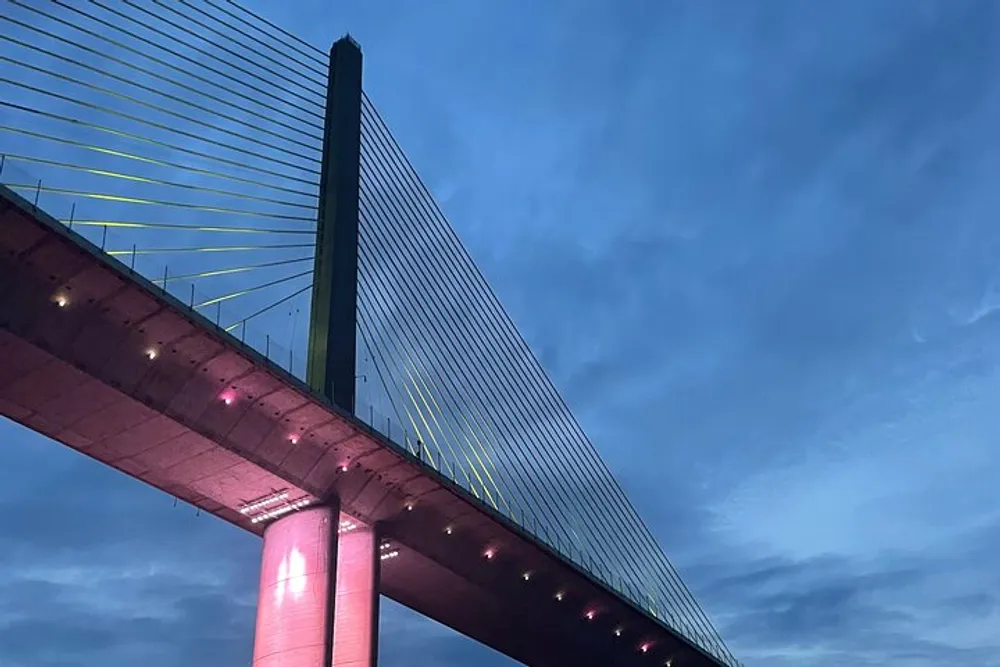 A modern suspension bridge illuminated with pink and yellow lights against a dusky blue sky