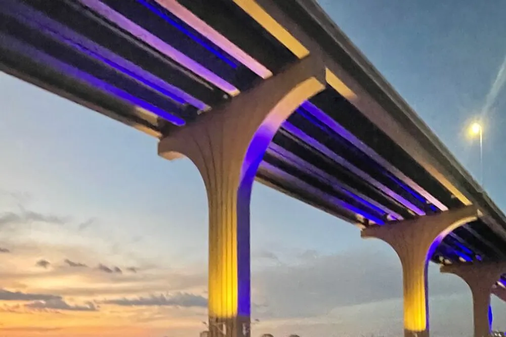 The photo shows a section of a bridge with illuminated blue lights lining its arches set against a softly lit evening sky