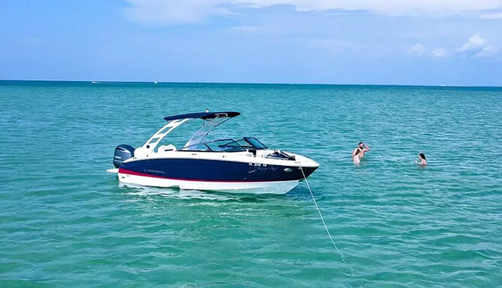 A motorboat is moored in clear turquoise waters while two people enjoy a swim nearby