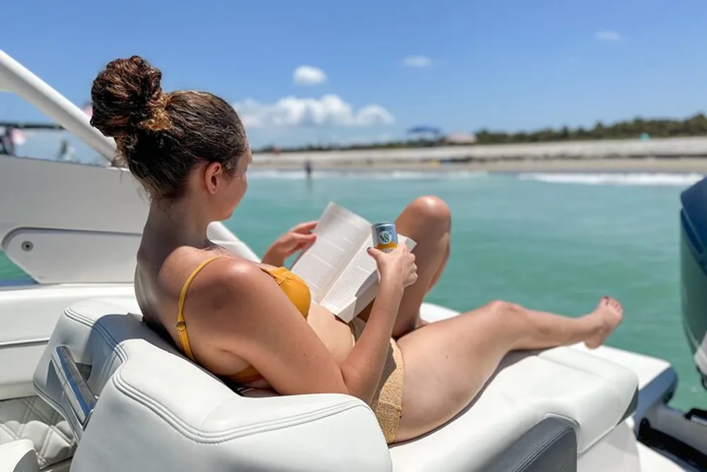 A person in a yellow bikini is relaxing on a boat reading a book with her legs propped up and a beverage can in hand against a backdrop of calm waters and a shoreline