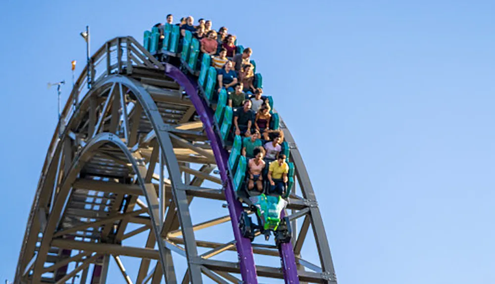 Thrill-seekers experience a moment of anticipation as they crest the peak of a roller coaster track against a clear blue sky