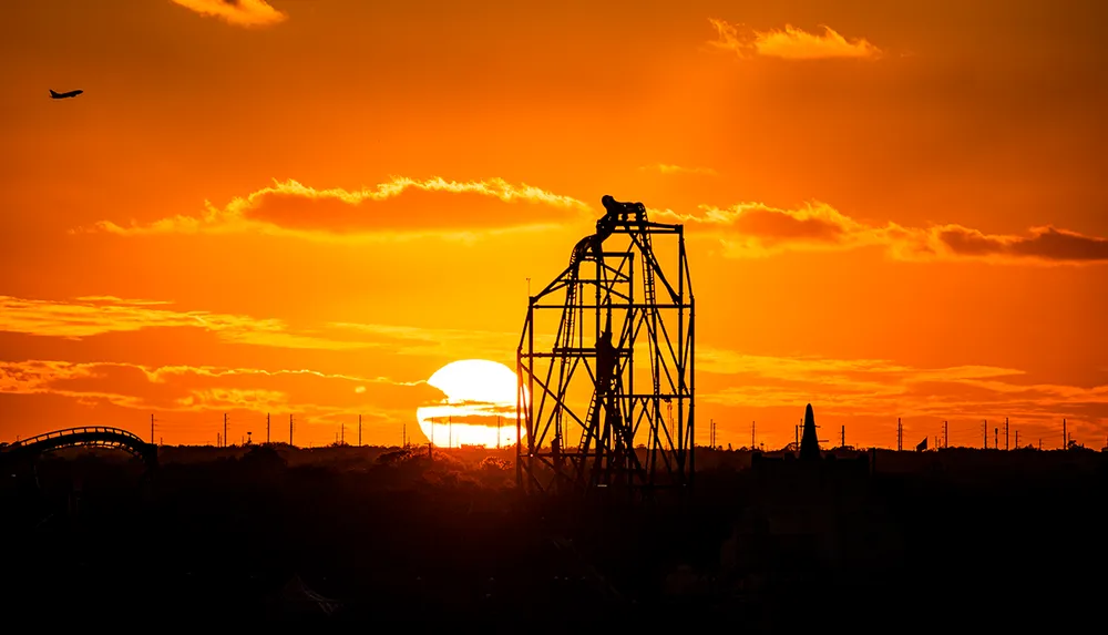 The image captures a roller coaster silhouette against a vibrant orange sunset sky with a plane flying in the distance