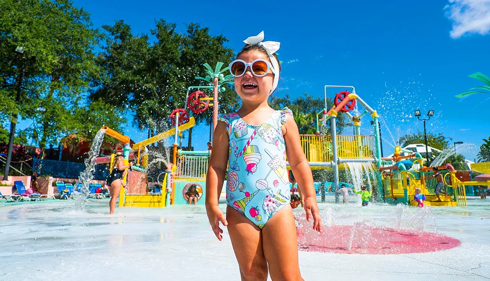 A joyful child in swimwear and sunglasses stands in a colorful splash pad area on a sunny day