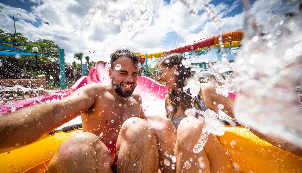 A man and a woman are joyfully riding down a water slide at a water park with splashes of water around them