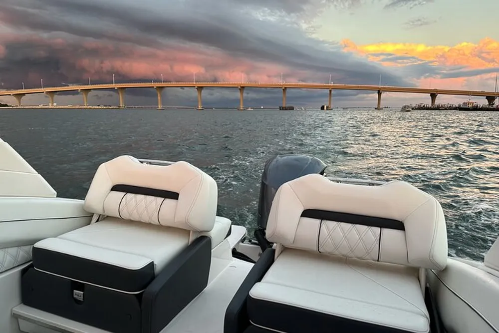 A view from a boat showing plush seats and an outboard motor with a scenic backdrop of a bridge and a dramatic sunset sky