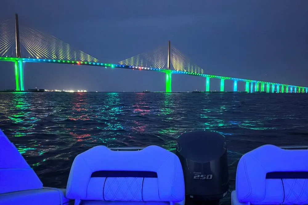 A brightly lit suspension bridge with blue and green lights reflects over the water at night viewed from the stern of a boat