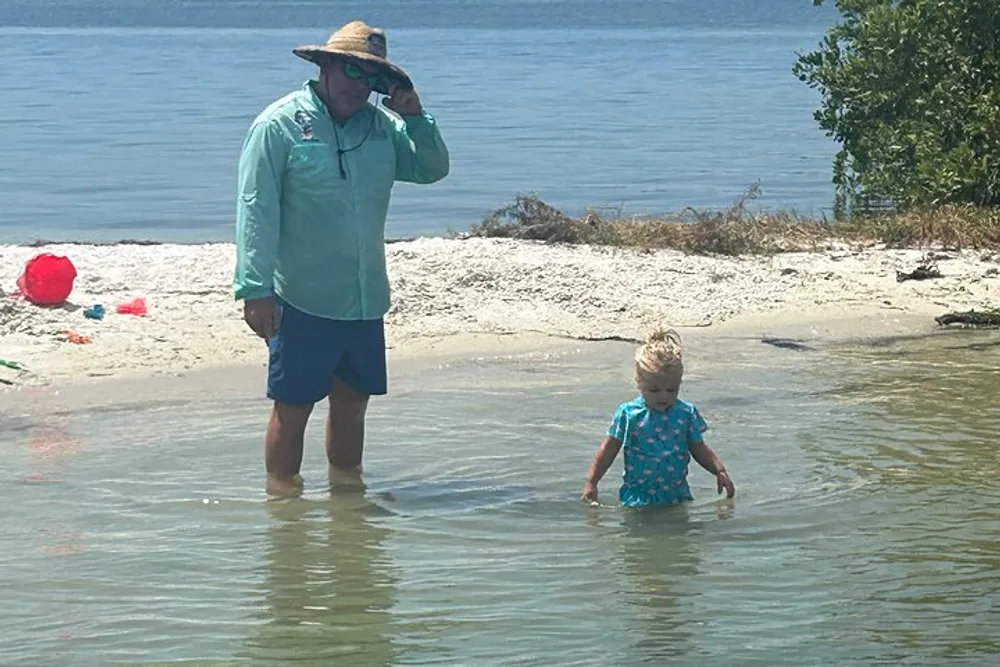 An adult and a child are standing in shallow water near the shore of a beach with the adult adjusting their hat and the child wading through the water