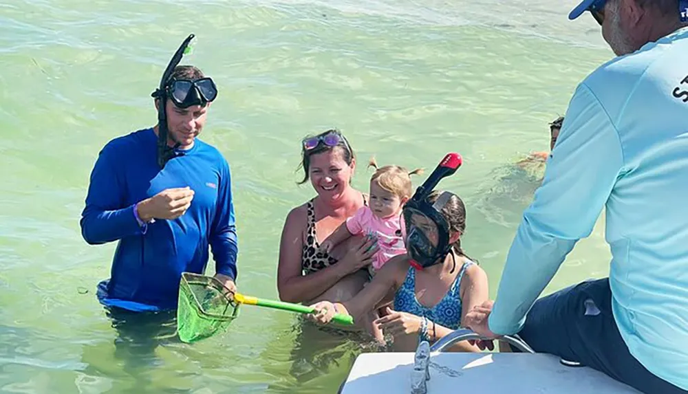 A family is enjoying a snorkeling activity in shallow waters with a guide showing them something while a woman holds a young child and another child wears snorkeling gear