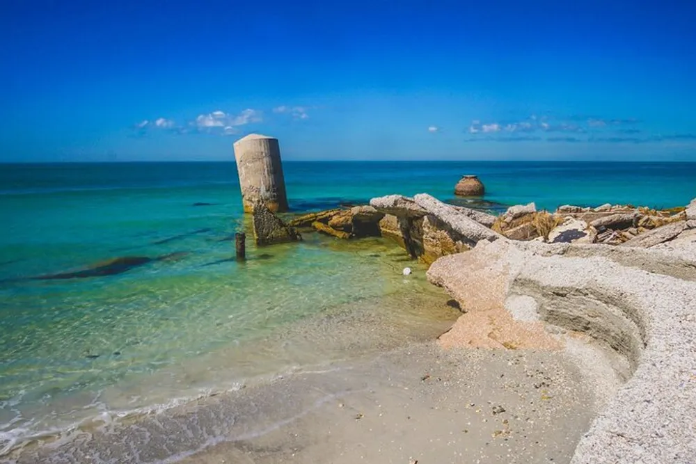 The image shows a serene beach with crystal-clear water broken concrete pillars and rocks that appear to be the remains of an old structure or jetty under a blue sky