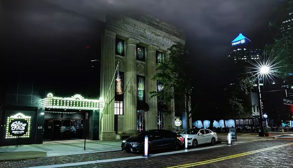 The image depicts a well-lit classic-style building at night showcasing an illuminated marquee and surrounded by modern cars and urban street elements