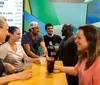 A group of people is gathered around a wooden table outdoors engaging in what appears to be a lively conversation over drinks and snacks at night