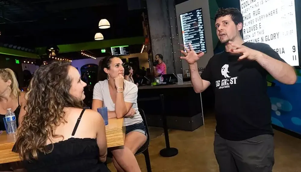 A man is animatedly speaking with his hands while two women listen attentively in what appears to be a casual social event at a bar or similar venue