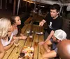 A group of people is gathered around a wooden table outdoors engaging in what appears to be a lively conversation over drinks and snacks at night