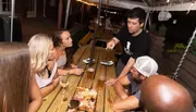 A group of people is gathered around a wooden table outdoors, engaging in what appears to be a lively conversation over drinks and snacks at night.