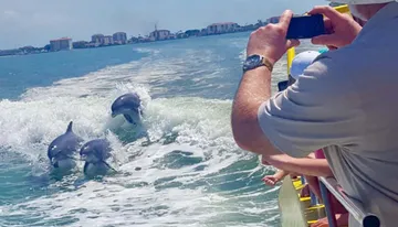 A person on a boat takes a photo of three dolphins jumping through the waves near the shore.