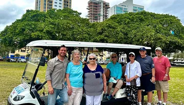 A group of seven people are smiling for a photo in front of a white open-air shuttle, with trees and a cityscape in the background.