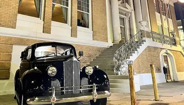 A vintage black car is parked in front of a building with grand steps and iron railings at night.