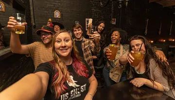 A group of cheerful people is taking a selfie in a bar, raising their drinks to the camera.