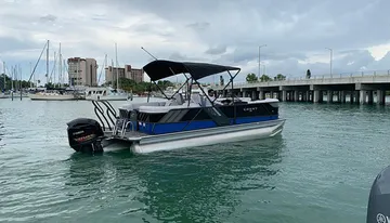 A pontoon boat with a black Bimini top is cruising near a bridge, with several docked sailboats and buildings in the background.