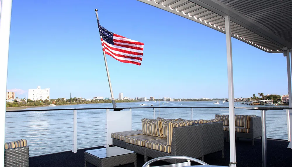 An American flag flies aboard a boat with comfortable seating overlooking a calm waterfront and a city skyline
