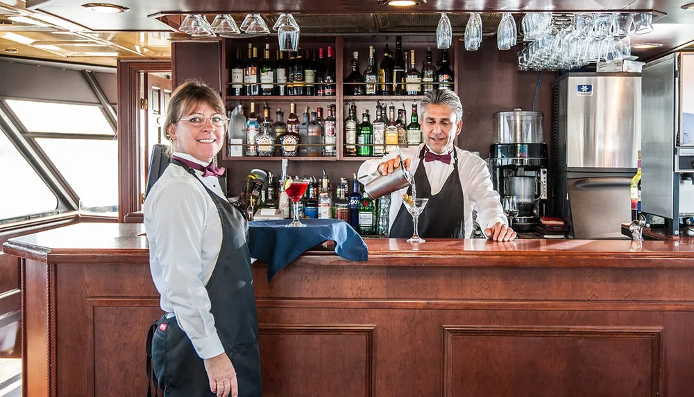 A bartender is pouring a drink beside a smiling colleague at a well-stocked bar
