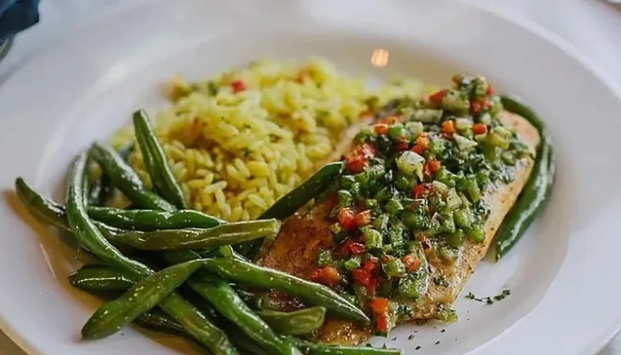 A plate of grilled fish topped with diced vegetables is served alongside seasoned yellow rice and green beans.