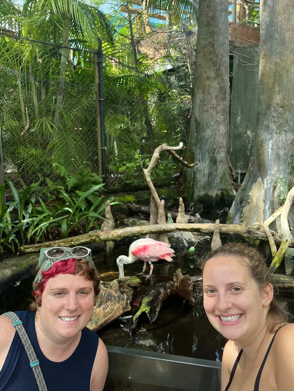 Two people are smiling for a selfie with a pink flamingo and lush greenery in the background