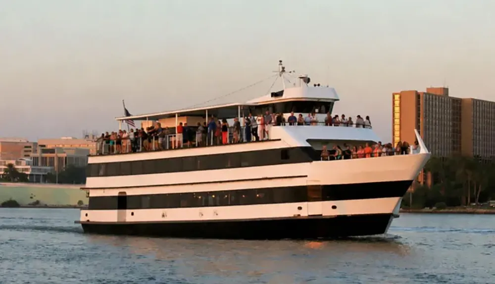 A large multi-deck boat filled with people is cruising on a body of water near a cityscape at sunset
