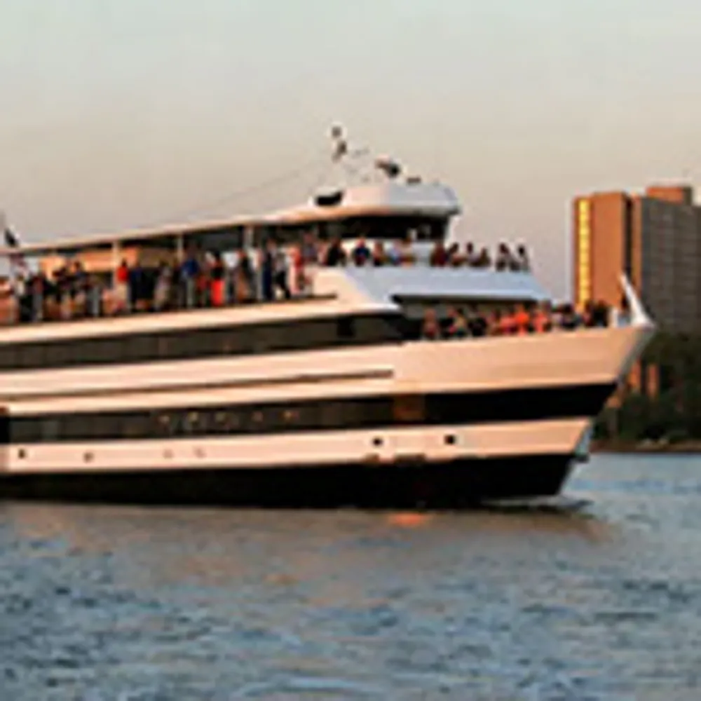 A large ferry with multiple decks is carrying people across the water near a shoreline with tall buildings at sunset