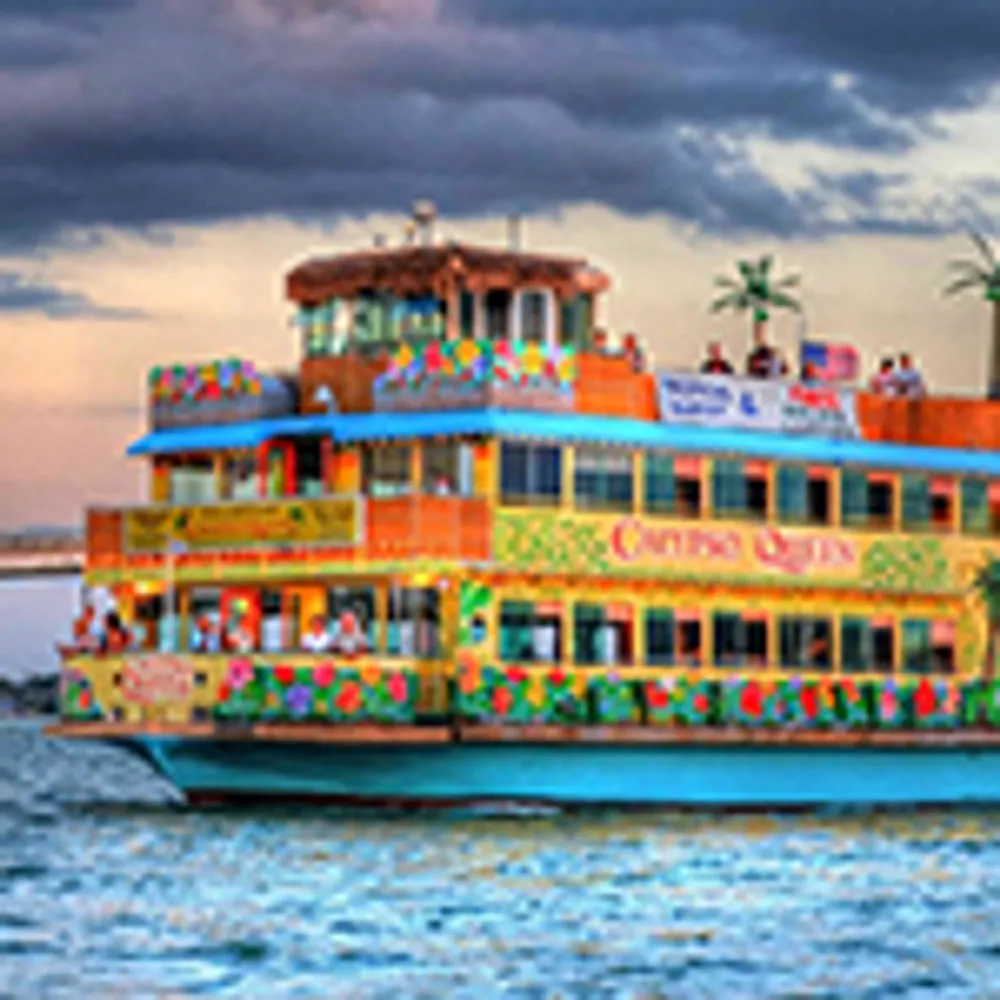 The image depicts a brightly colored festive boat with people on board sailing on a body of water under a cloudy sky