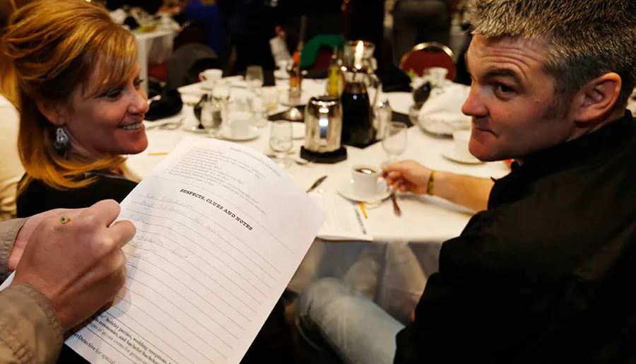A woman and a man are looking at a document titled Suspect Clues and Notes together at a banquet table with empty dishes and coffee cups.