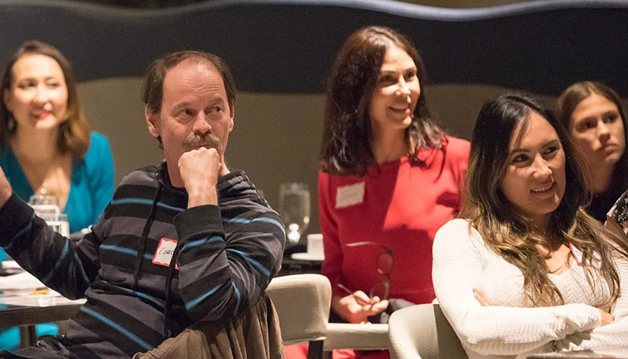 A group of people are engaged in what appears to be an attentively listening audience at an event, with some smiling and one man resting his chin on his hand.