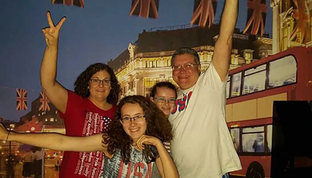 A happy family of four is posing with raised hands in front of a backdrop featuring British-themed elements including the Union Jack and a red double-decker bus